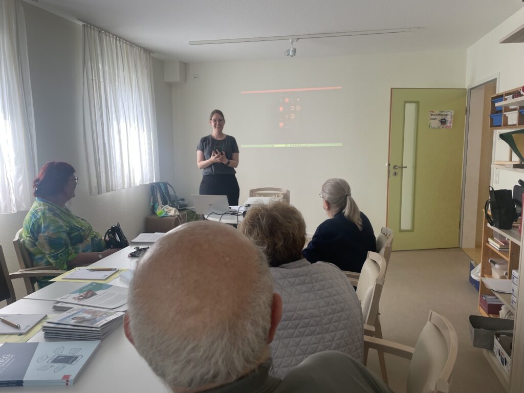 Ein Raum mit einem Tisch an dessen Ende Martina steht. Eine Gruppe älterer Menschen sitzt am Tisch und schaut Martina an.