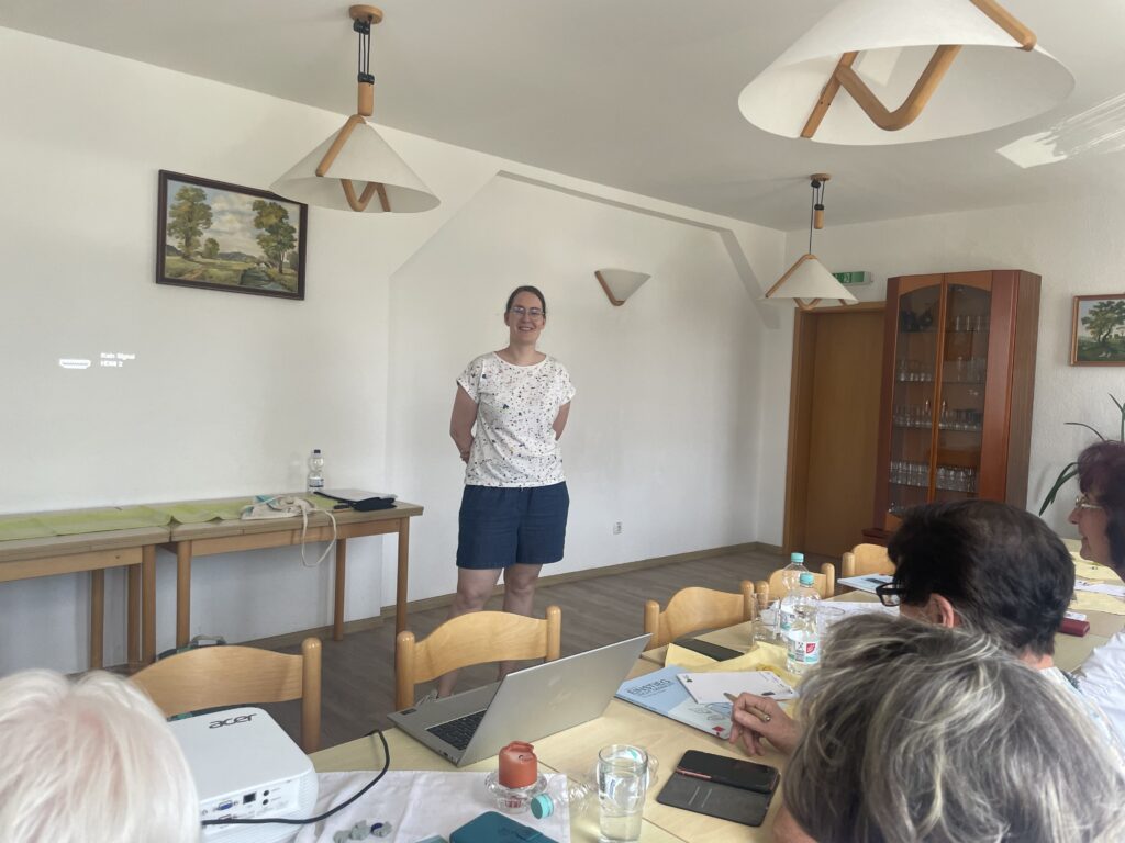 Martina steht vor einer Gruppe älterer Menschen, die an einem Tisch sitzen. Sie spricht mit ihnen.