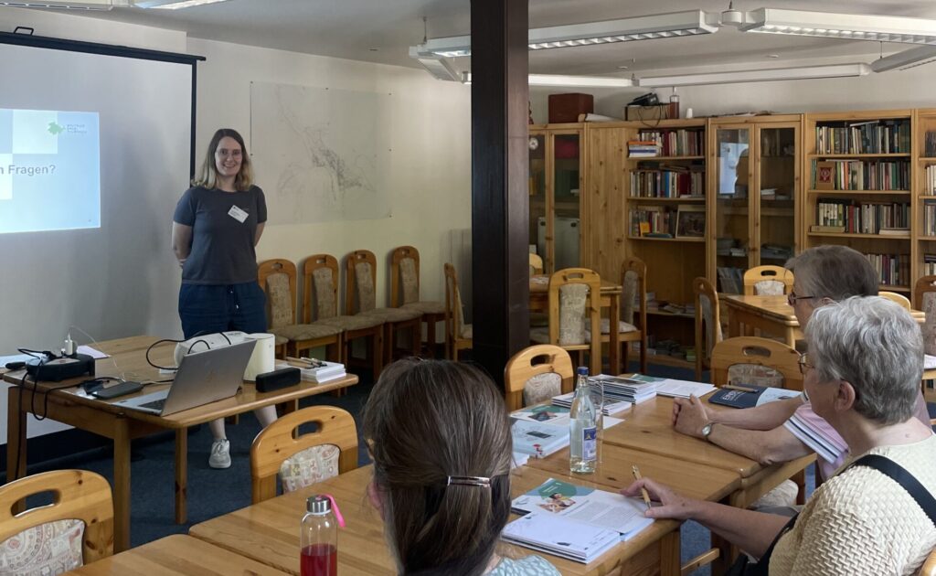 Martina steht vor einer Gruppe älterer Menschen, die an einem Tisch sitzen. Hinter ihr ist eine Präsentation an die Wand geworfen, Sie lächelt in die Kamera.