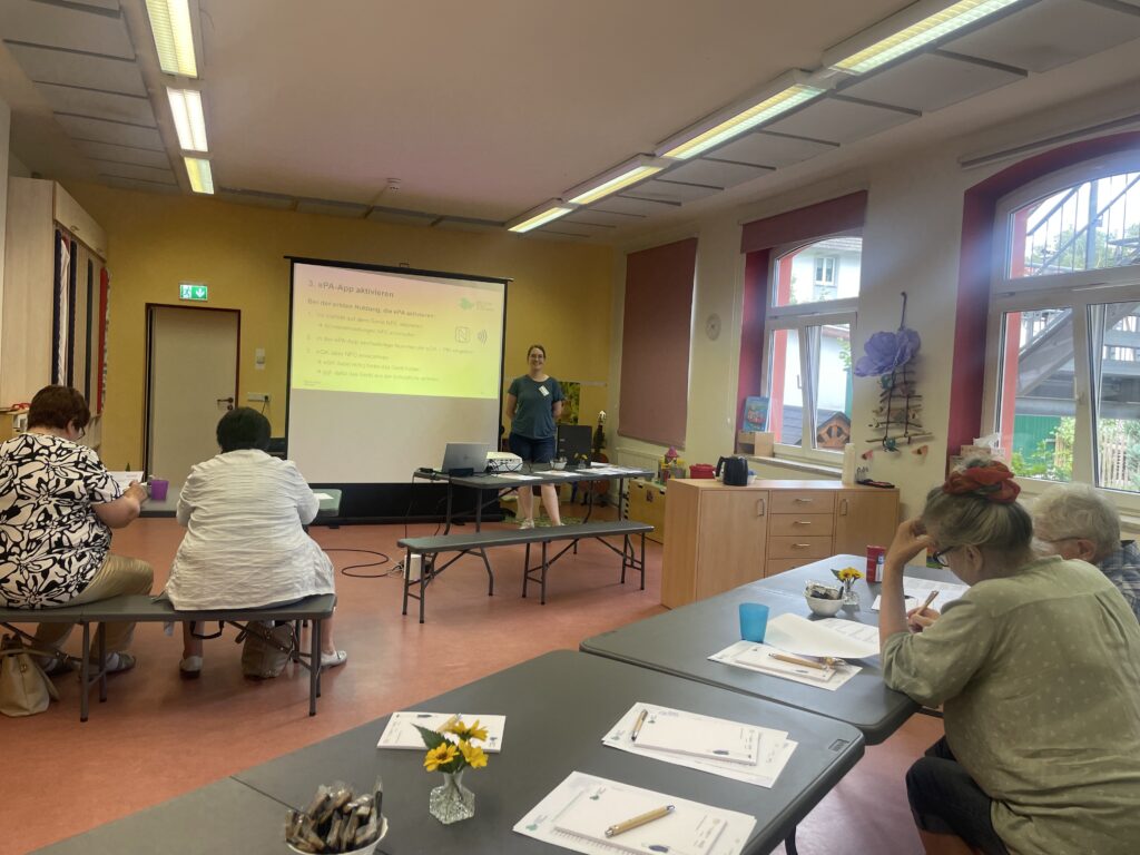 Ein großer Raum mit im Raum verteilten Tischen, an denen zum Teil ältere Menschen sitzen. Vor ihnen steht Martina vor einer Präsentation und lächelt in die Kamera.