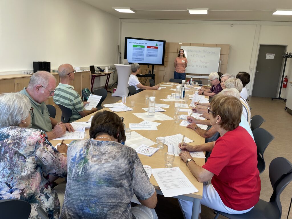 Sophie steht vor einer Gruppe älterer Menschen, die an einem langen Tisch sitzen.