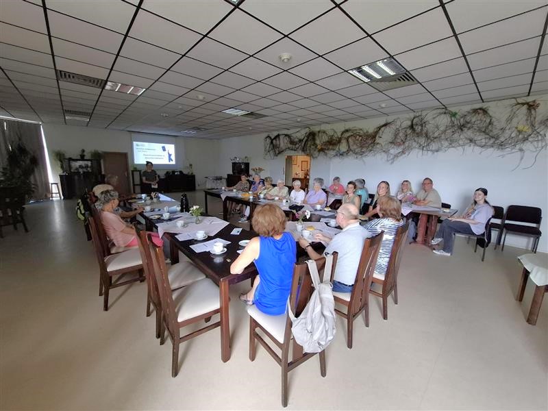 Ein dunkler Raum mit einer großen Sitzgruppe älterer Menschen. Ganz vorne im Raum ist Sophie beim Halten eines Vortrags zu sehen.