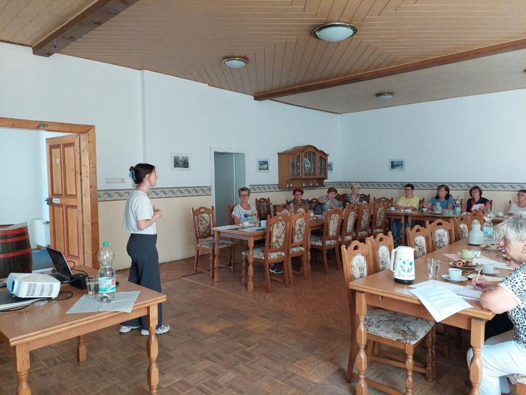 Sophie steht vor einer großen Gruppe älterer Menschen, die in einem großen Saal auf Holzstühlen sitzen.