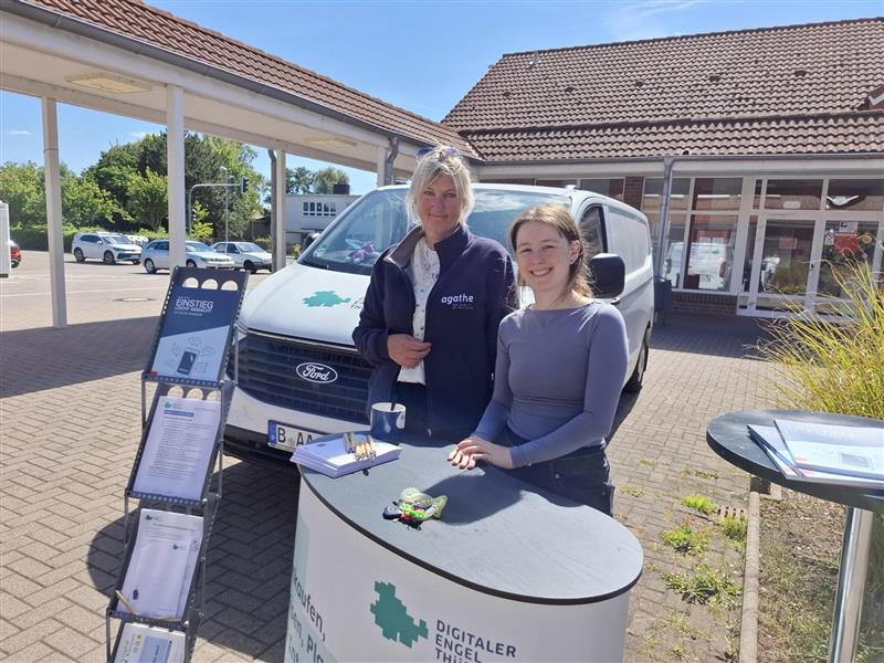 Sophie steht mit einer Person am Stand des Digitalen Engels Thüringen, der draußen aufgebaut ist. Beide lächeln in die Kamera.. Hinter ihnen ist das Infomobil zu sehen.