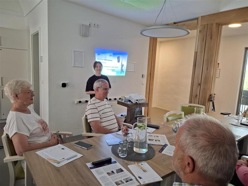 Sophie steht vor einer Gruppe älterer Menschen, die an einem runden Tisch sitzt. Auf dem Tisch sind Zettel und Gläser verteilt. Sophie lächelt in die Kamera.