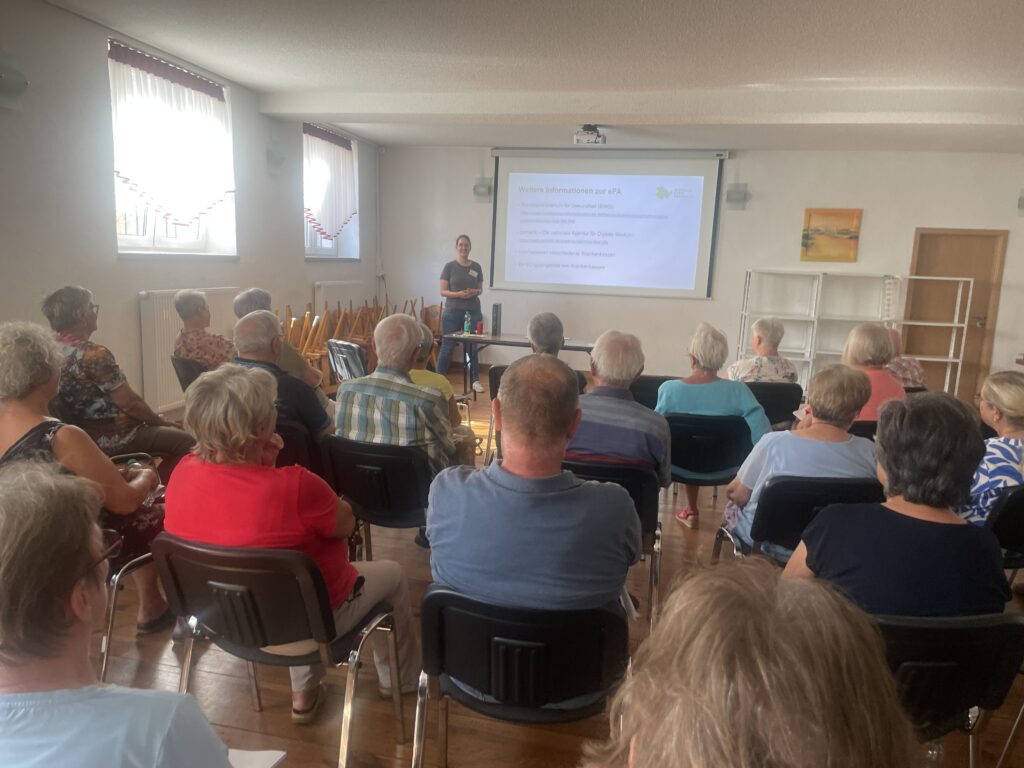 Martina steht vor einer Gruppe älterer Menschen und lächelt in die Kamera. Im Hintergrund sieht man eine Leinwand mit Martinas Präsentation.