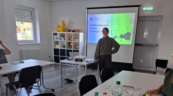 Sophie steht vor einer Leinwand, auf die eine Präsentation projiziert wird. Sie lächelt in die Kamera.