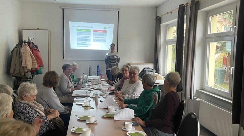 Person steht vor Leinwand mit Präsentation, mehrere Personen sitzen an langem Tisch mit Tassen, Tellern und Unterlagen in hellem Raum mit Fenstern