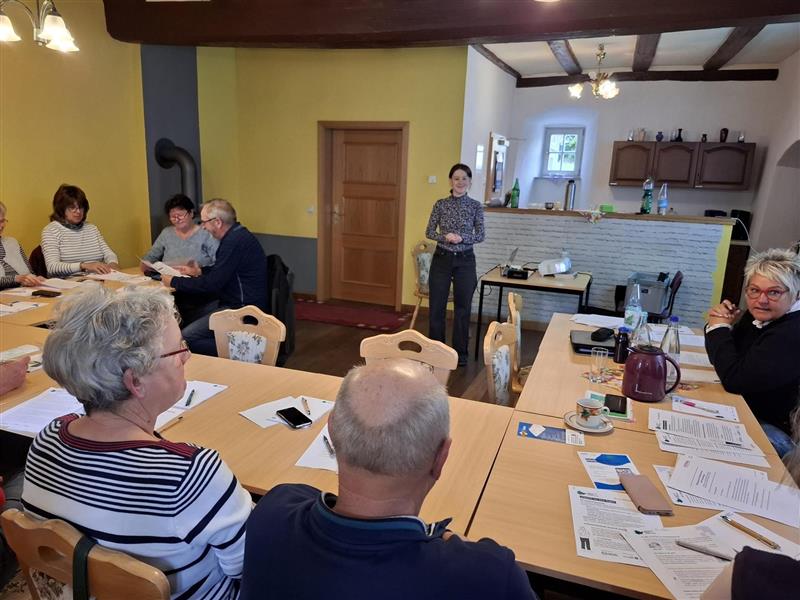 Person steht vor einem Tisch mit mehreren Personen, die an Papieren und Dokumenten sitzen, in einem Raum mit gelben Wänden und Holzmöbeln.