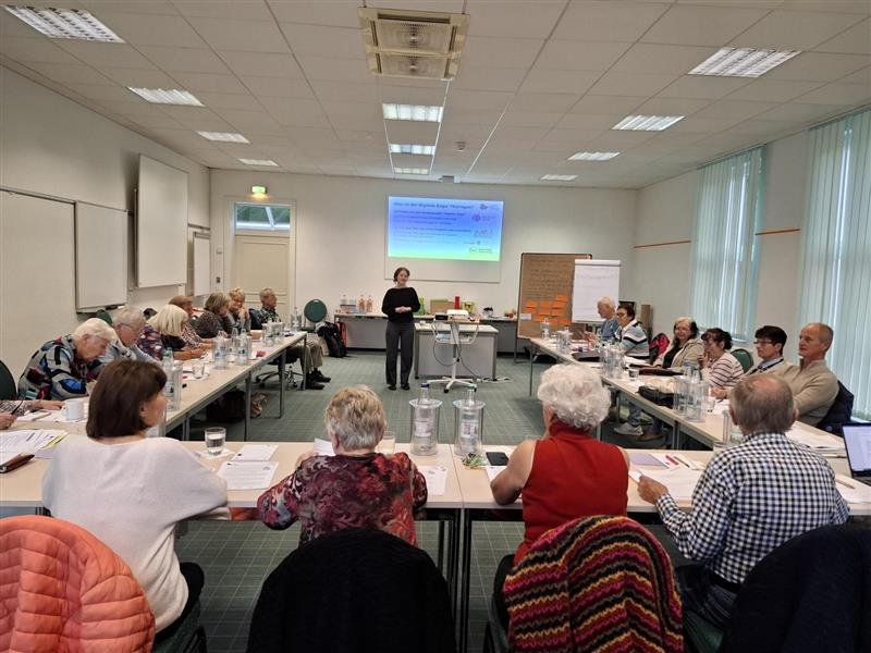 Senior:innen sitzen an einer Tischgruppe in U-Form. In der Mitte steht Sophie und spricht zu ihnen.