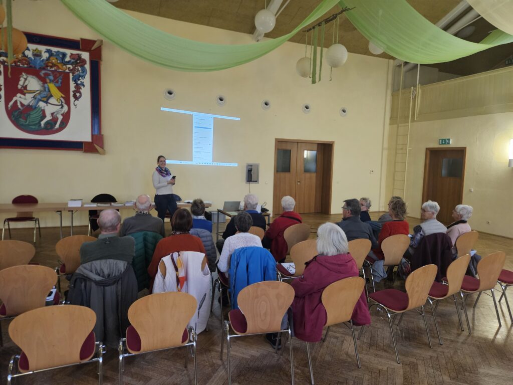 Person steht vor einer Gruppe sitzender Menschen in einem Saal mit Holzboden und grünen Stoffbahnen an der Decke.