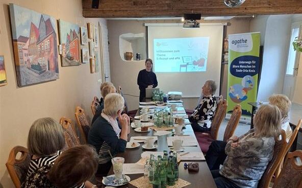 Mehrere ältere Frauen sitzen an einem langen Tisch in einem Raum mit Holzbalken und betrachten eine Präsentation an der Wand.