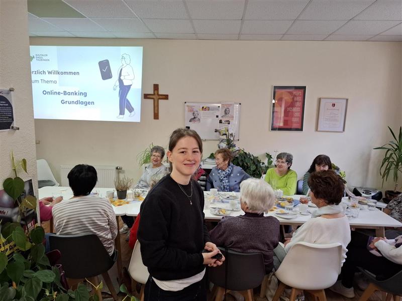 Mehrere Personen sitzen an einem Tisch in einem Raum mit einem Kreuz an der Wand und einer Projektion mit dem Text 'Herzlich Willkommen zum Thema Online-Banking Grundlagen'.