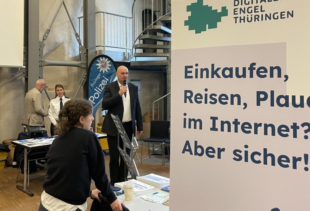 Wendeltreppe mit Metallgeländer in einem Raum mit hohen Wänden, davor ein Banner mit dem Text 'Digitaler Engel Thüringen Einkaufen, Reisen, Plaudern im Internet? Aber sicher!' und Personen an einem Infostand.