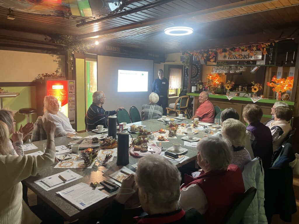 Person steht vor einem Bildschirm mit Präsentation in einem Raum mit herbstlicher Dekoration, mehrere Personen sitzen an einem Tisch mit Unterlagen und Getränken.