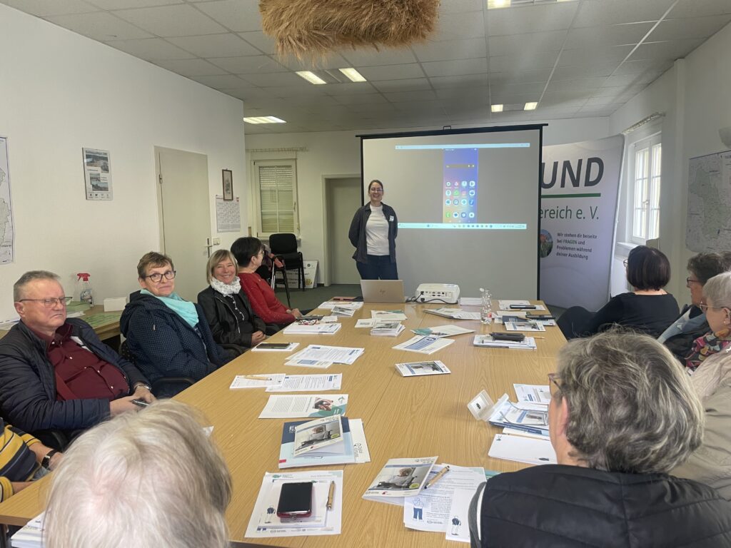 Person steht vor einem Beamerbildschirm mit Smartphone-Startbildschirm, mehrere Personen sitzen an einem Tisch mit Unterlagen in einem Besprechungsraum.