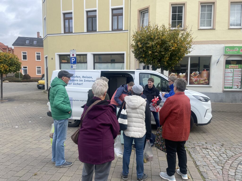 Gruppe älterer Menschen steht vor einem weißen Lieferwagen mit Text auf der Seite auf einem gepflasterten Platz in einer Kleinstadt.