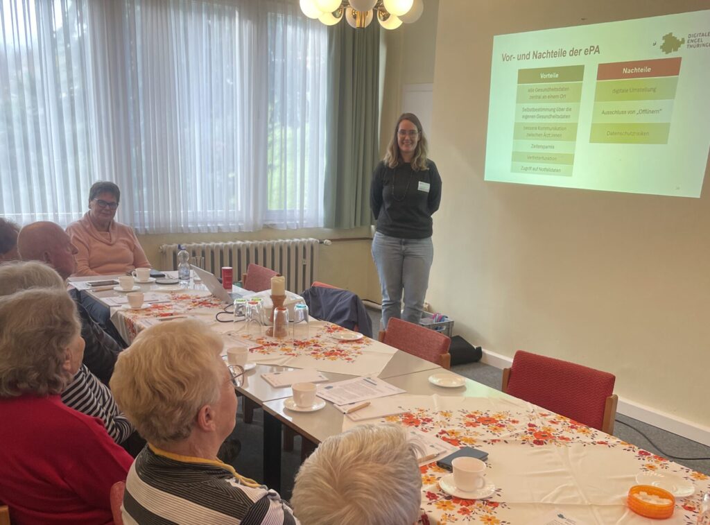 Frau steht vor einer Wand mit projizierter Präsentation, mehrere ältere Personen sitzen an einem mit Blumen-Tischdecke bedeckten Tisch mit Tassen und Unterlagen.