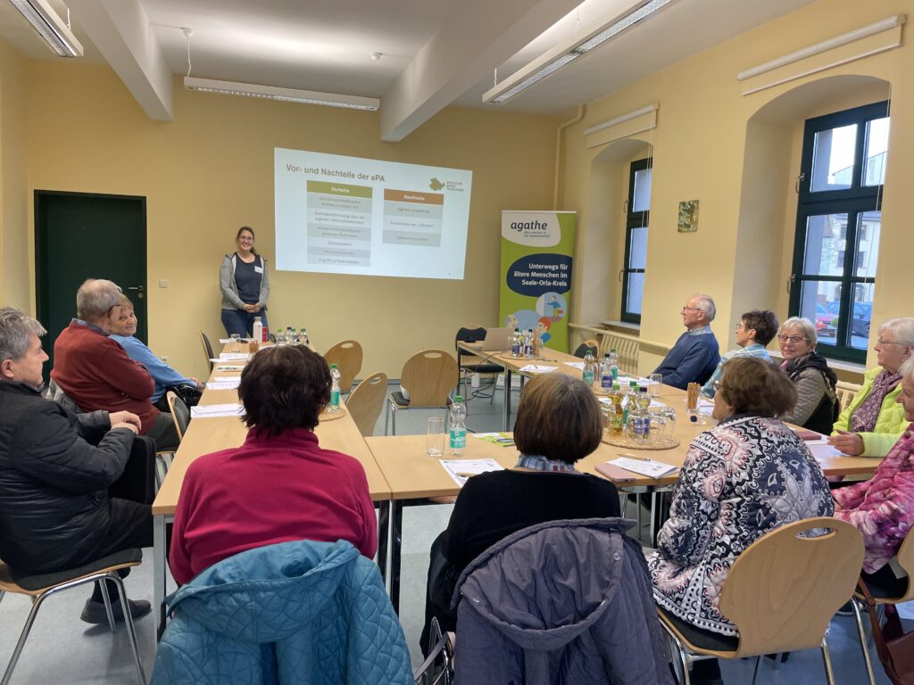 Mehrere Personen sitzen an Tischen in einem hellen Raum und hören einer stehenden Vortragenden zu, die vor einer Präsentationstafel steht.