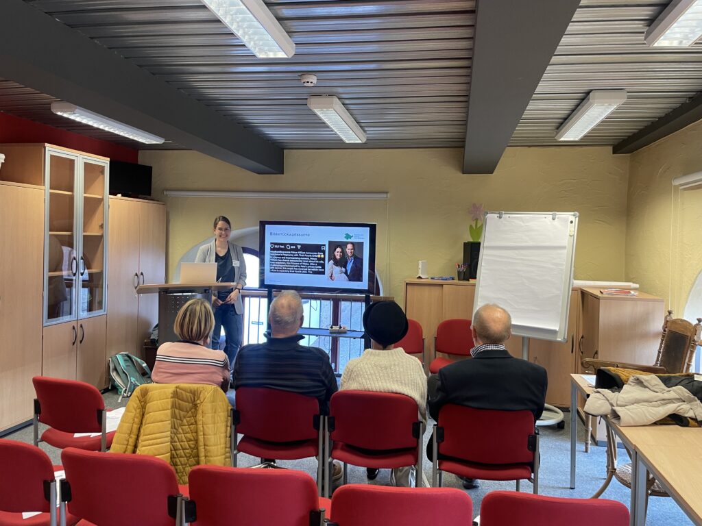 Person steht vor Bildschirm mit Präsentation, vier Personen sitzen auf roten Stühlen und hören zu, im Raum Schrank, Flipchart und Tische.