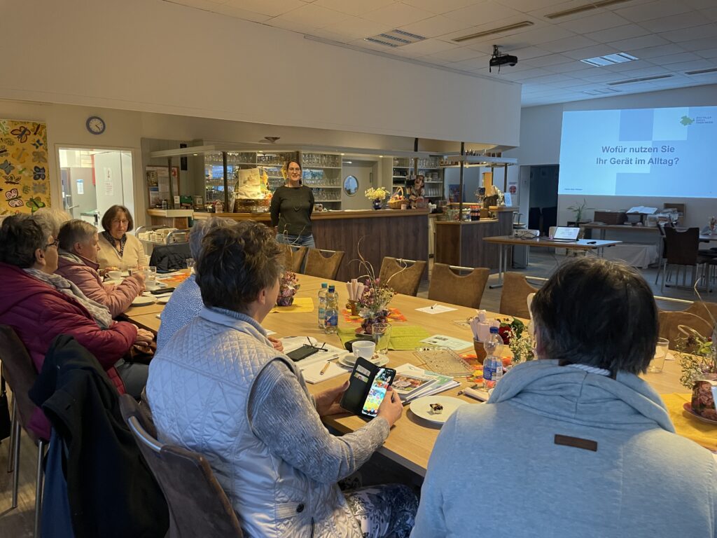 Mehrere Personen sitzen an einem langen Holztisch in einem Raum mit Theke, eine Person hält ein Smartphone, im Hintergrund eine Projektion mit dem Text 'Wofür nutzen Sie Ihr Gerät im Alltag?'.