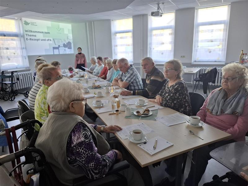Gruppe älterer Menschen sitzt an einem langen Tisch in einem hellen Raum, vorne steht eine Vortragende neben einer Präsentation mit dem Text 'Willkommen zum Thema E-Rezept und ePA'.