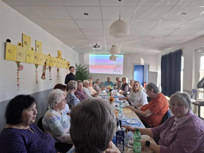 Mehrere ältere Personen sitzen an einem langen Tisch in einem hellen Raum, an der Wand hängen gelbe Bilder mit Gesichtern, vorne steht eine Person und präsentiert vor einer Leinwand.
