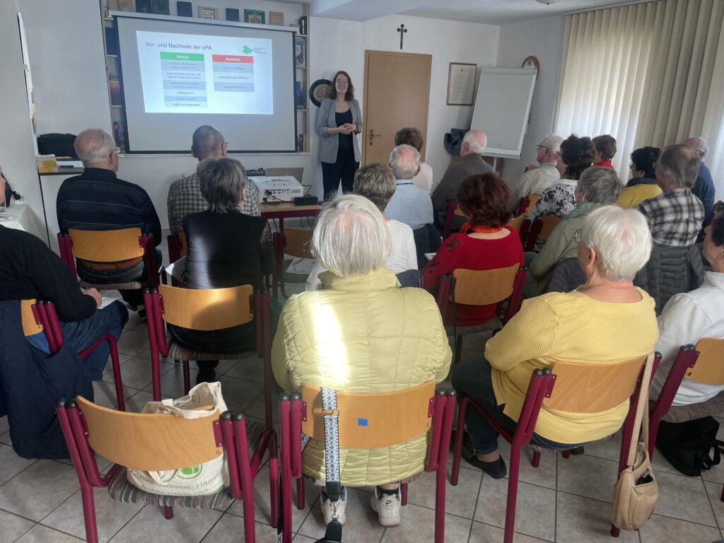 Mehrere ältere Personen sitzen auf Stühlen in einem Raum und hören einer Frau zu, die vor einer Leinwand mit einer Präsentation steht.