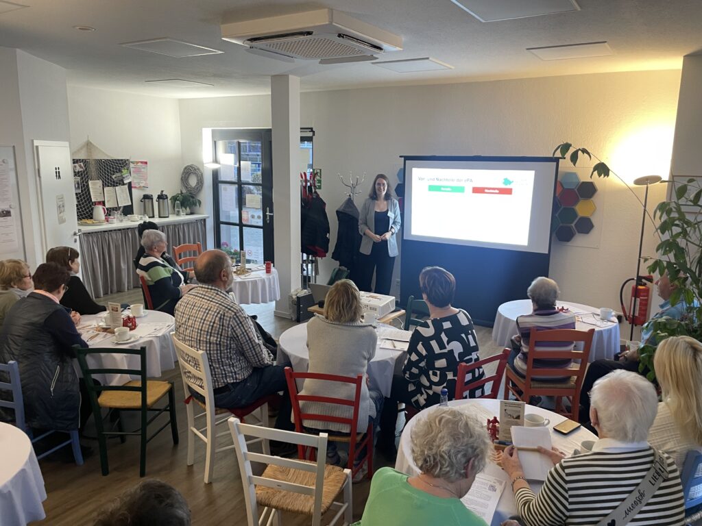 Mehrere ältere Personen sitzen an Tischen in einem Raum und schauen auf eine Leinwand, auf der eine Präsentation gezeigt wird.
