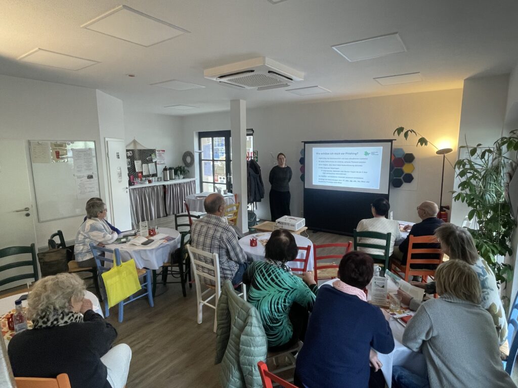 Mehrere ältere Personen sitzen an Tischen in einem Raum und schauen auf eine Leinwand, auf der eine Präsentation gezeigt wird.