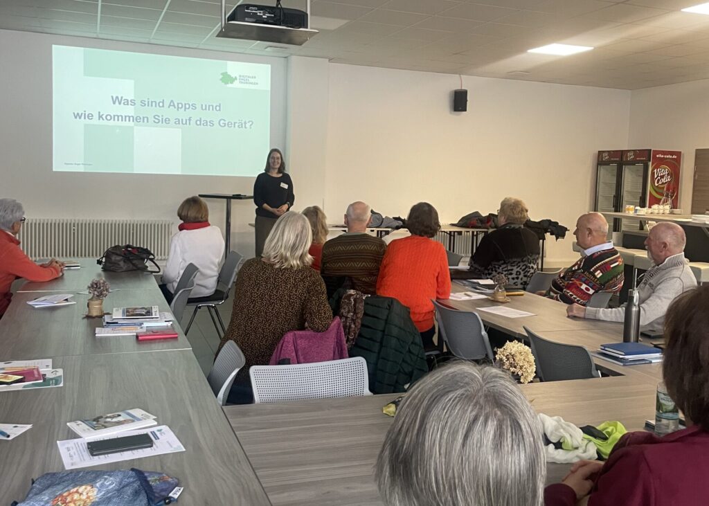 Mehrere ältere Menschen sitzen an Tischen in einem hellen Raum und hören einer Frau zu, die vor einer Leinwand mit dem Text 'Was sind Apps und wie kommen Sie auf das Gerät?' steht.