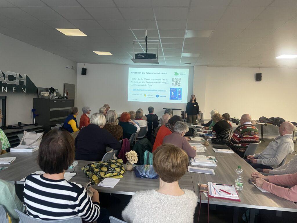 Mehrere Personen sitzen an Tischen in einem hellen Seminarraum und hören einer stehenden Frau zu, die vor einer Projektionsleinwand spricht.