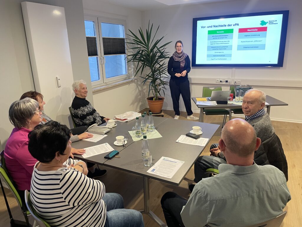 Mehrere Personen sitzen an einem Tisch in einem Besprechungsraum, eine Frau steht vor einem Bildschirm mit einer Präsentation.