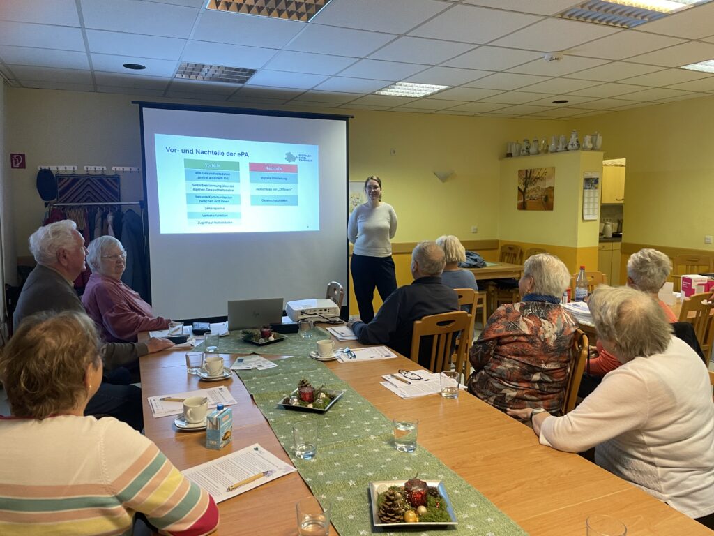 Person steht vor einer Gruppe älterer Menschen, die an einem Tisch sitzen und eine Präsentation mit Text und Diagrammen auf einer Leinwand ansehen.