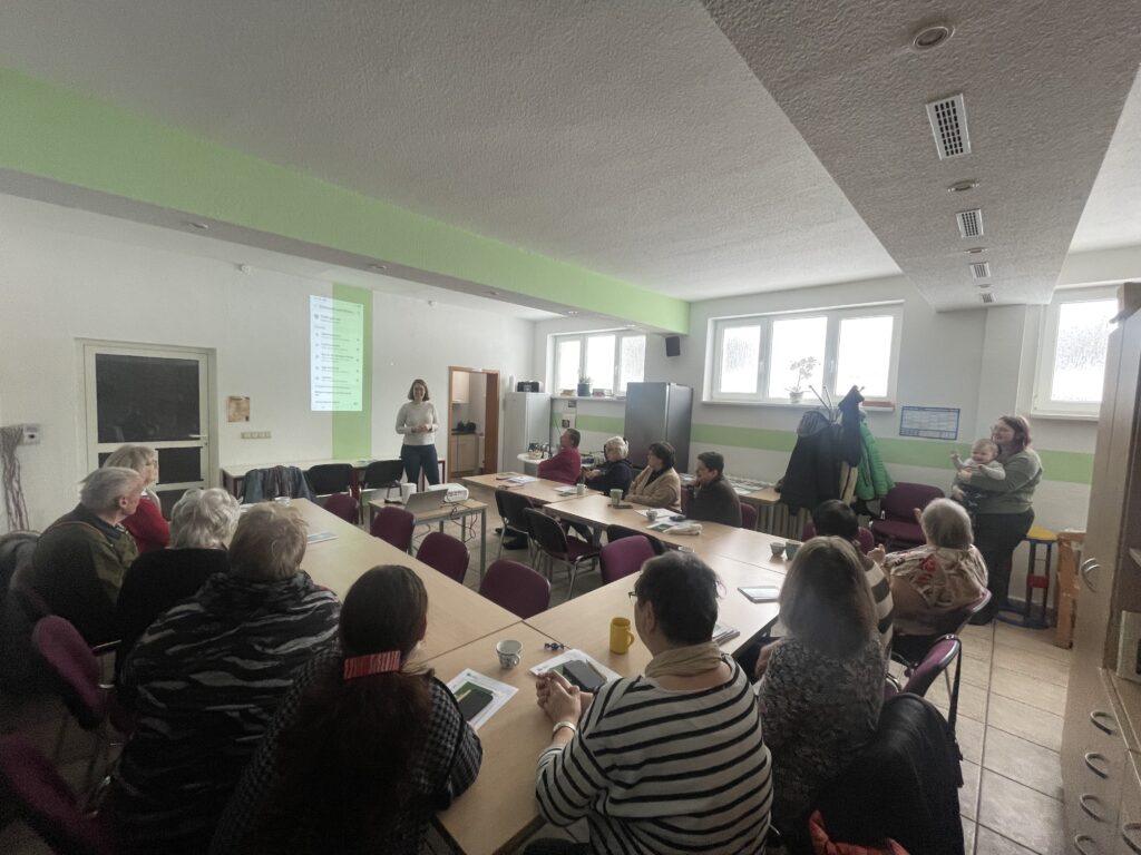 Mehrere ältere Personen sitzen an Tischen in einem hellen Raum und hören einer stehenden Frau zu, die vor einer projizierten Präsentation spricht.