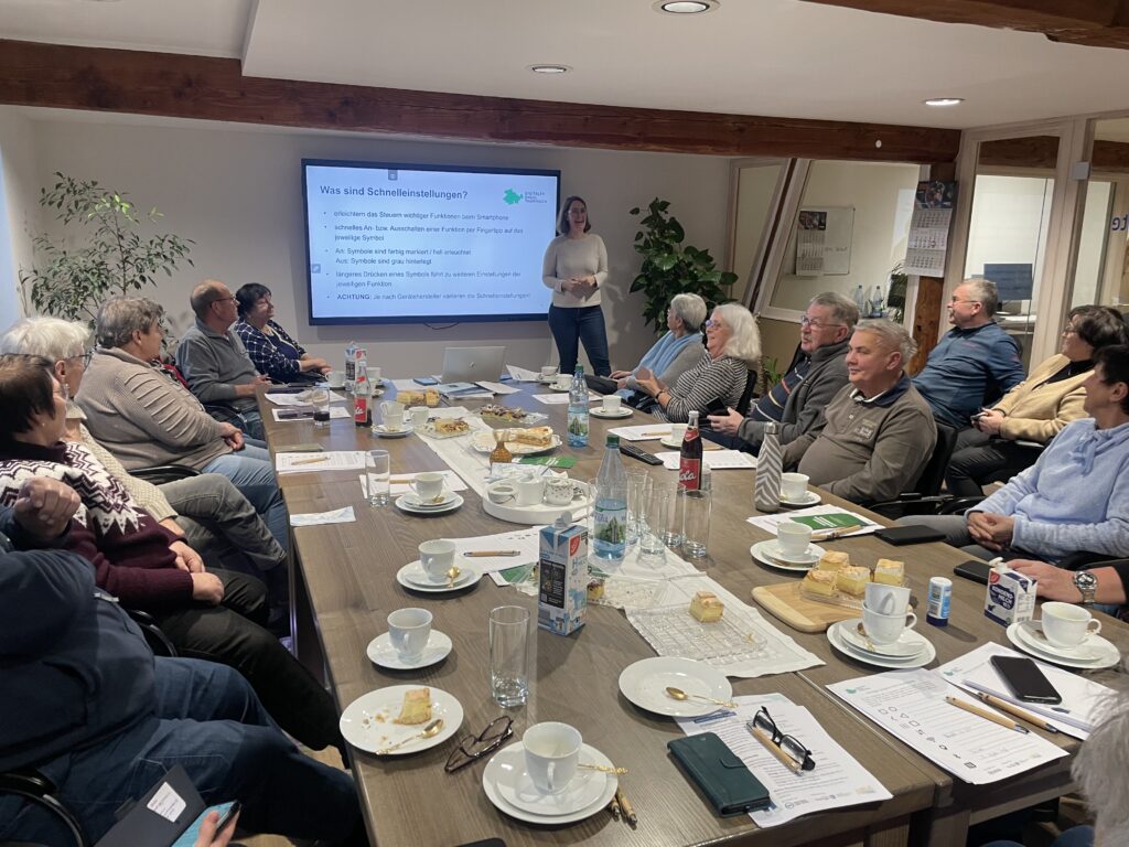 Person steht vor großem Bildschirm mit Präsentationstext, umgeben von mehreren sitzenden Personen an einem langen Tisch mit Kaffeetassen, Getränken und Snacks