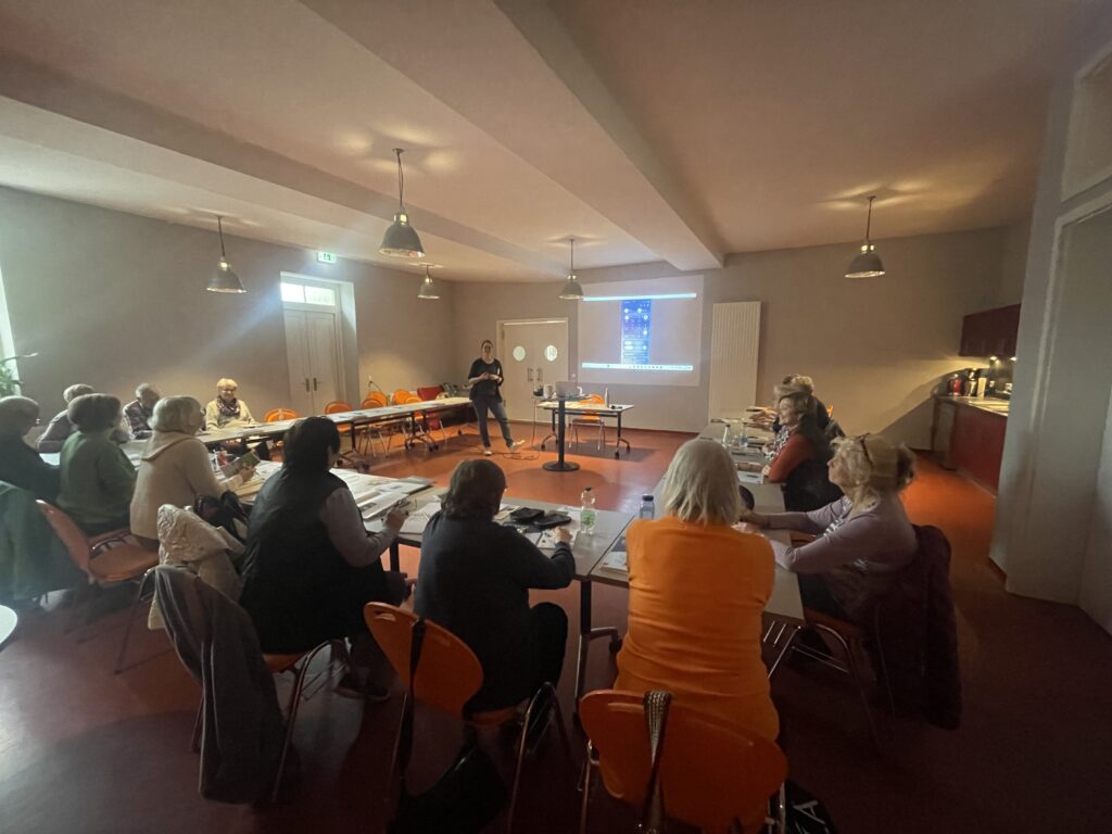 Martina steht vor einer Gruppe älter Menschen, die in einer U-Form vor ihr sitzt.