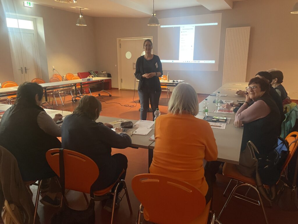 Martina steht vor einer Gruppe älter Menschen, die in einer U-Form vor ihr sitzt.