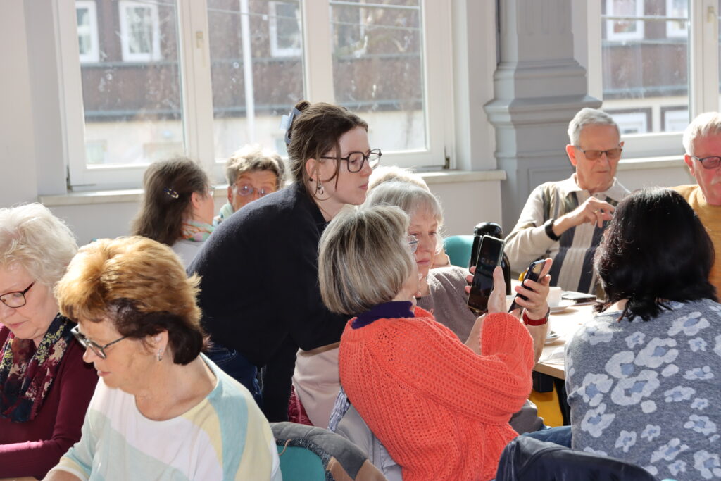 Sophie steht in einer Gruppe älterer Menschen und schaut auf ein Handy, das ihr hingehalten wird.