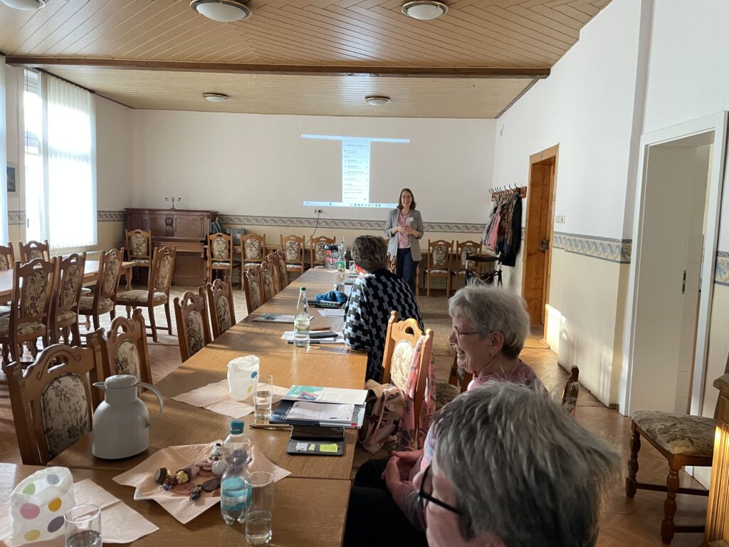 Mehrere Personen sitzen an einem langen Holztisch in einem hellen Raum mit Holzdecke, eine Frau steht vor einer projizierten Präsentation an der Wand
