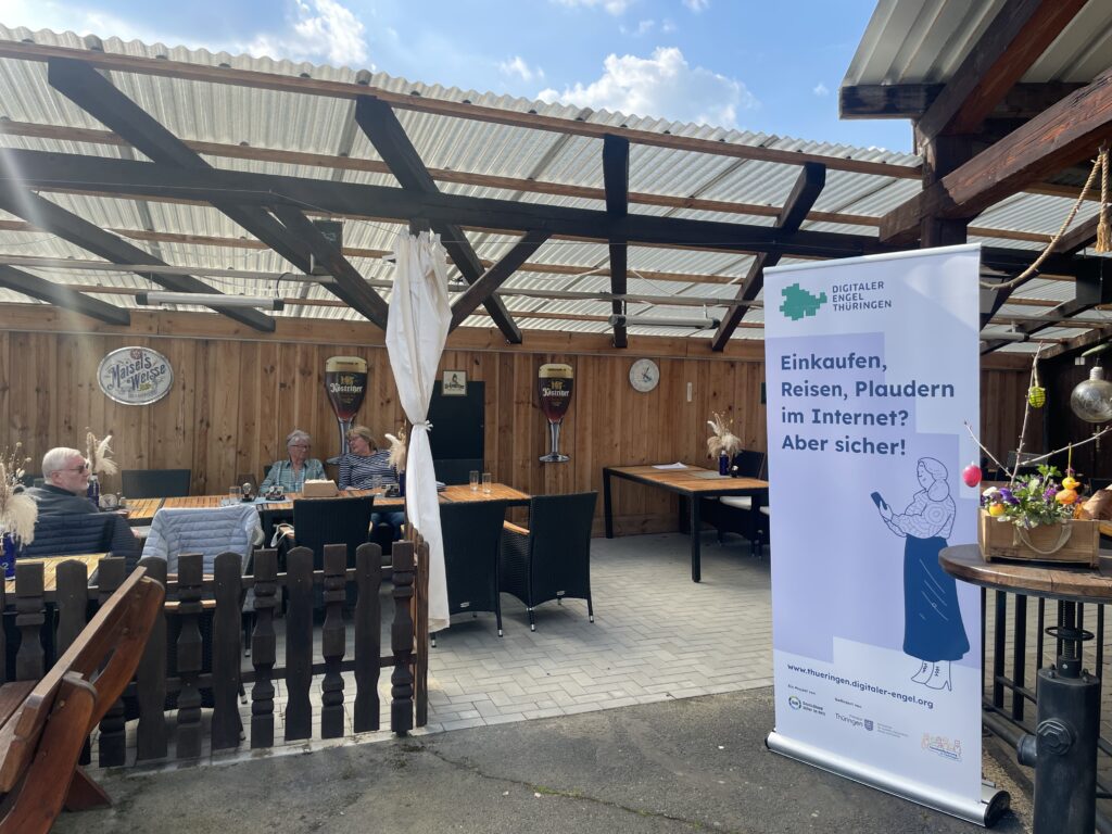 Überdachter Außenbereich mit Holzwänden, mehreren Personen an Tischen und einem aufgestellten Roll-up mit der Aufschrift 'Einkaufen, Reisen, Plaudern im Internet? Aber sicher!'