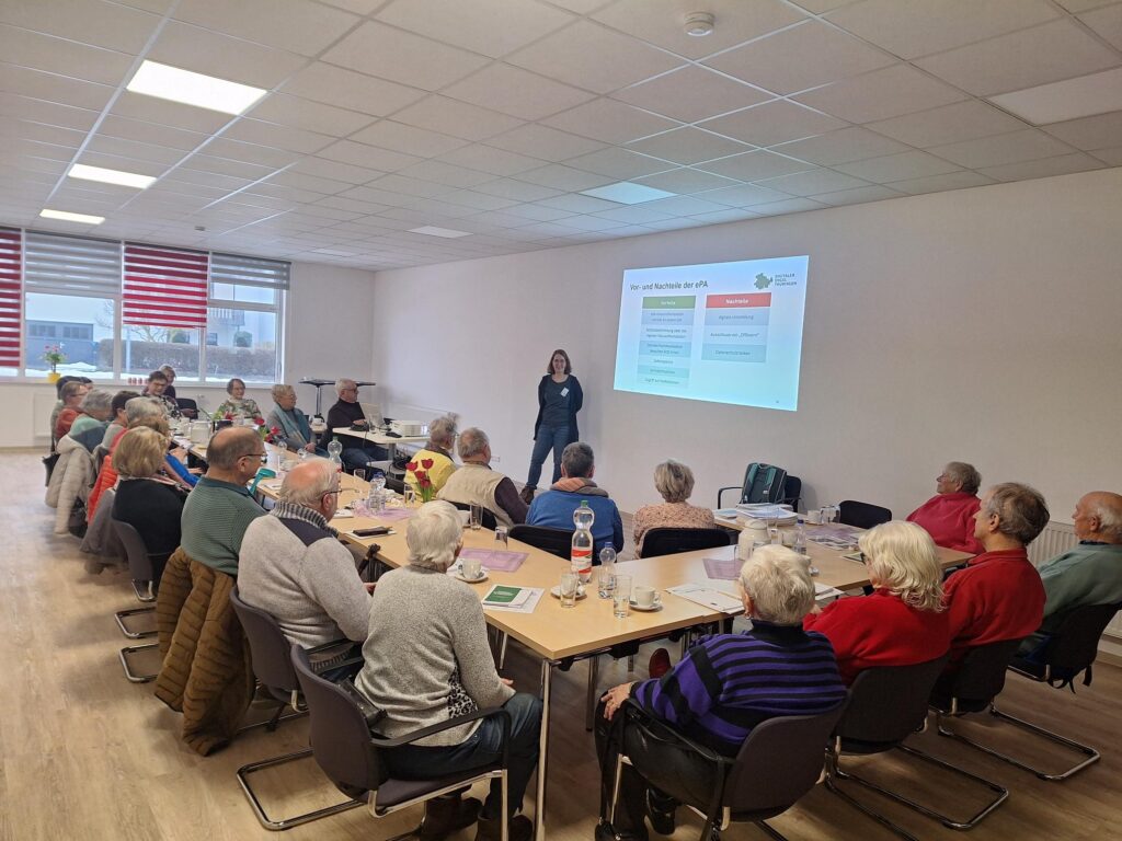 Gruppe älterer Menschen sitzt an Tischen in einem hellen Raum und hört einer Frau zu, die vor einer Wand mit einer Präsentation steht