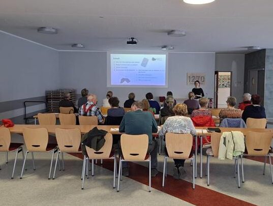 Gruppe von Menschen sitzt in einem hellen Raum an Tischen und schaut auf eine Präsentation an der Wand