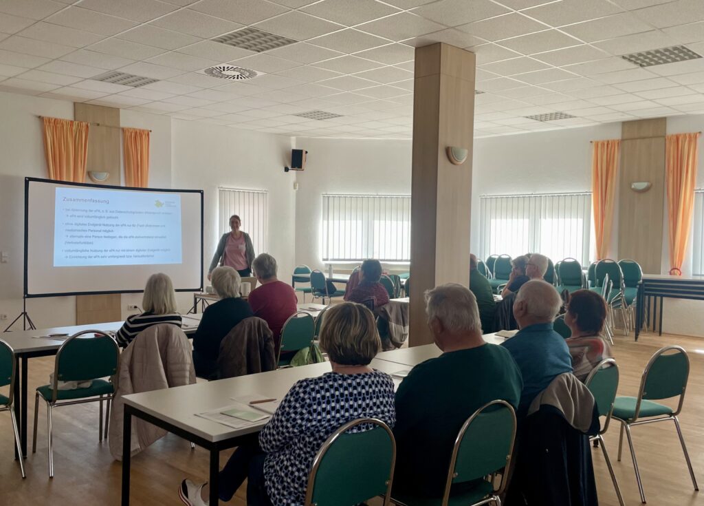 Mehrere ältere Personen sitzen in einem hellen Seminarraum an Tischen und hören einer stehenden Frau zu, die vor einer Leinwand mit Text präsentiert
