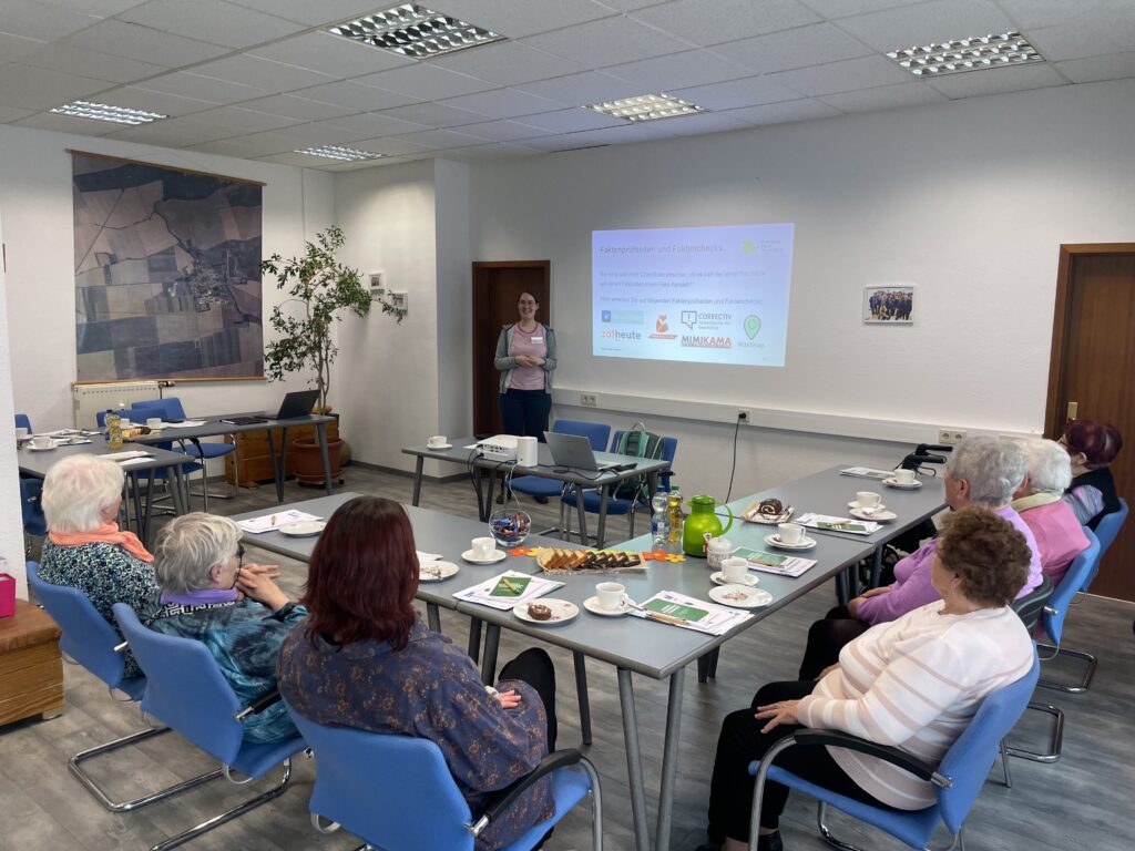 Gruppe älterer Menschen sitzt an Tischen in U-Form und hört einer stehenden Person zu, die vor einer Wand mit projizierter Präsentation spricht