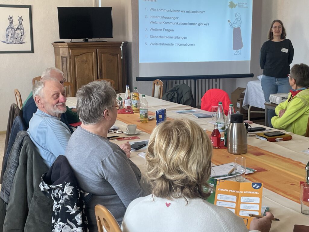 Martina steht vor einer Leinwand, an der eine Präsentation projiziert wurde. Vor ihr sitzen ältere Menschen an einem Tisch und schauen zu ihr.