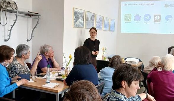 Frau steht vor einer Gruppe älterer Menschen in einem hellen Raum mit Wandbildern und projizierter Präsentation