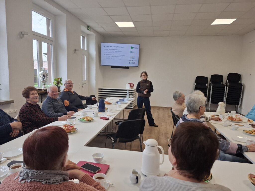 Sophie steht in der Mitte einer Gruppe älterer Menschen und lächelt in die Kamera. Auf Tischen sind Informationsmaterialen ausgelegt. Hinter Sophie ist eine Leinwand aufgebaut.