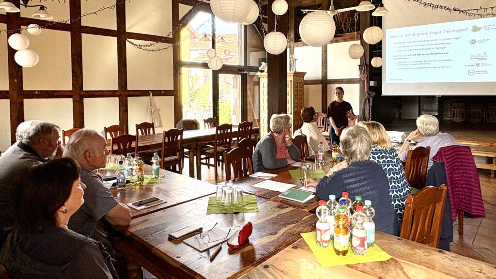 Personengruppe sitzt an langen Holztischen in einem Fachwerkraum und hört einer Vortragenden zu, die vor einer Leinwand steht