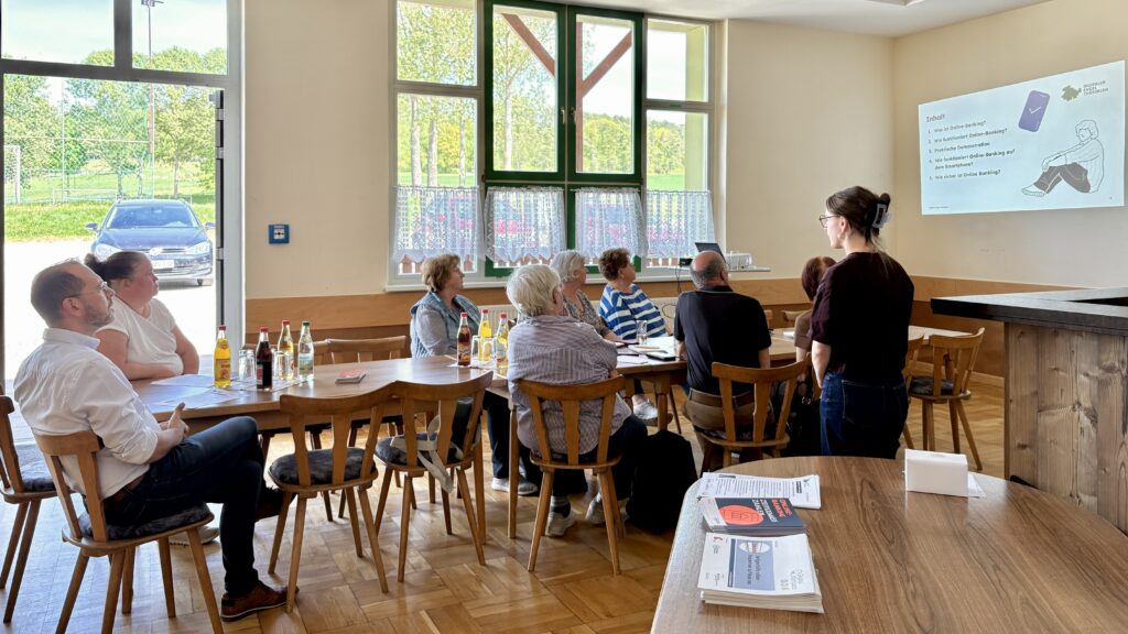 Sophie steht vor einer Gruppe älterer Menschen, die an einem langen Tisch sitzen. Im Hintergrund ist eine Präsentation auf die Wand projiziert.